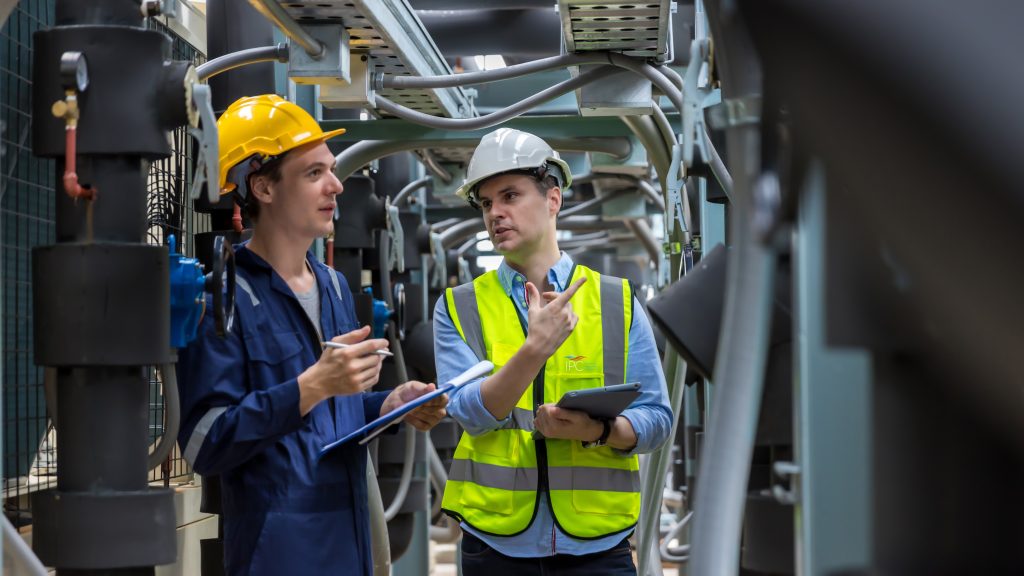 Two men holding clipboards and a tablet inspecting an area with pipes and electrical systems