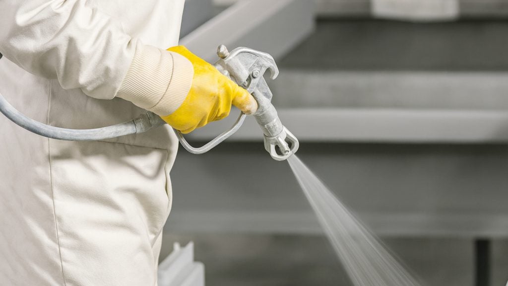 Close-up of worker in white protective suit and yellow gloves applying fireproof paint to stop fire on steel beam.