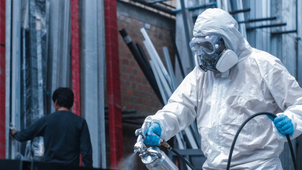 Two workers in white protective suits, one in the foreground applying fireproof paint to stop fire on steel beam., another in the background