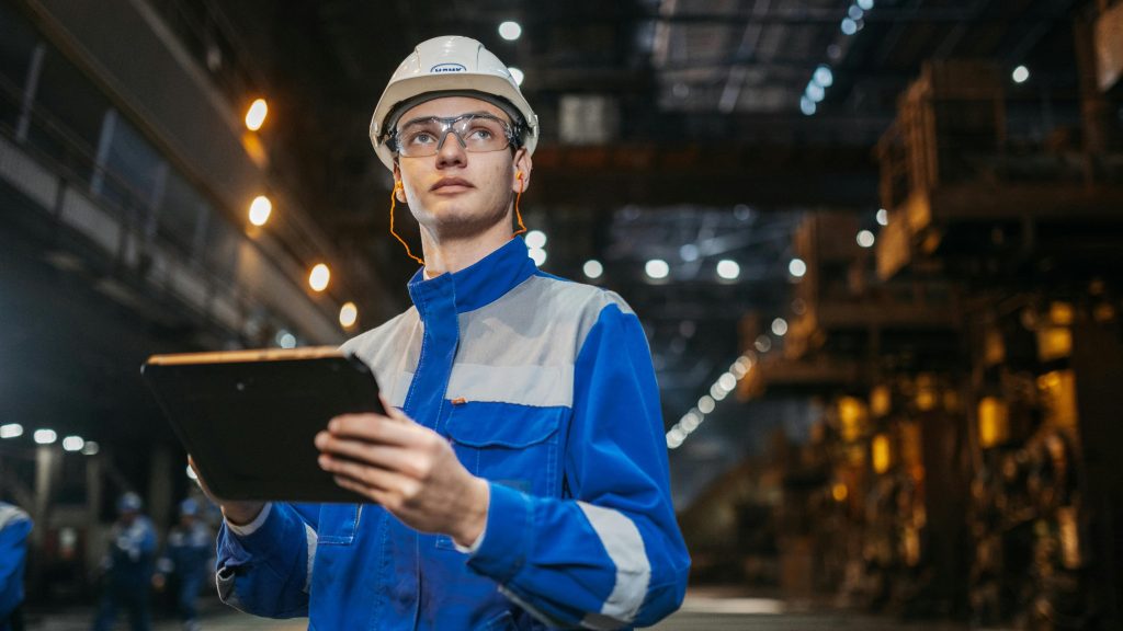 Man in hi-vis jacket, helmet, and goggles holding a clipboard in a warehouse