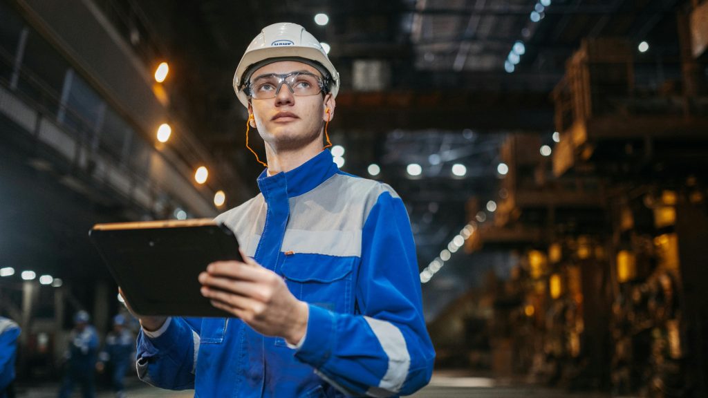 Man in hi-vis jacket, helmet, and goggles holding a clipboard in a warehouse