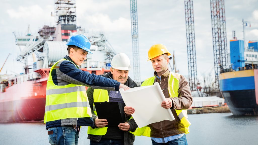 Three men in hi-vis jackets reviewing documents with boats in the background